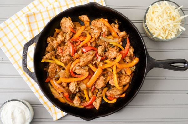 A cast iron skillet, filled with chicken breast pieces, sliced bell pepper, and sliced onion. Resting on a tea towel and a grey table. A bowl of sour cream in the bottom left corner, and a bowl of shredded cheese in the top right corner.