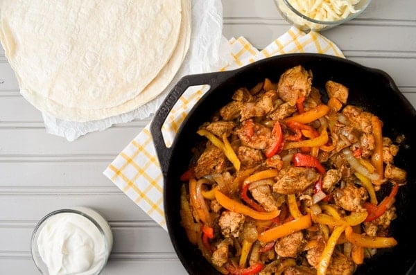 A cast iron skillet is filled with chicken breasts, sliced peppers, and onions. On the table next to it rests a bowl of sour cream, shredded cheese, and flour tortillas.