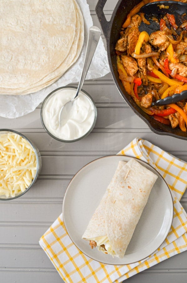 A grey wood table with all of the fixings for chicken fajitas, including shredded cheese, sour cream, and tortillas. A cast iron skillet is in the top right corner, containing chicken fajitas.