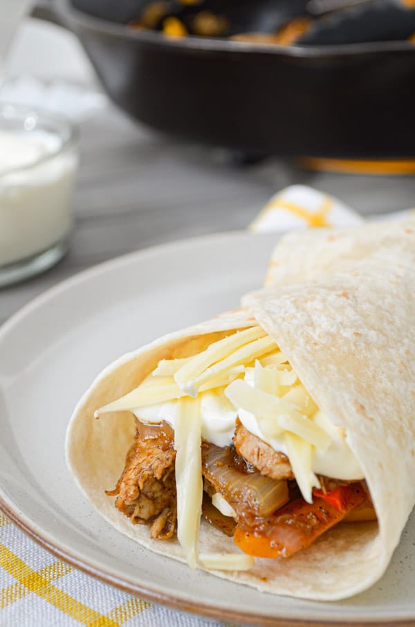 A chicken fajita rests on a plate. With a skillet of chicken fajitas in the background.
